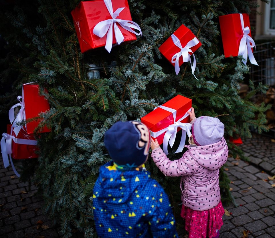 Zwei Kinder hängen rote Päckchen an den Geschenkebaum